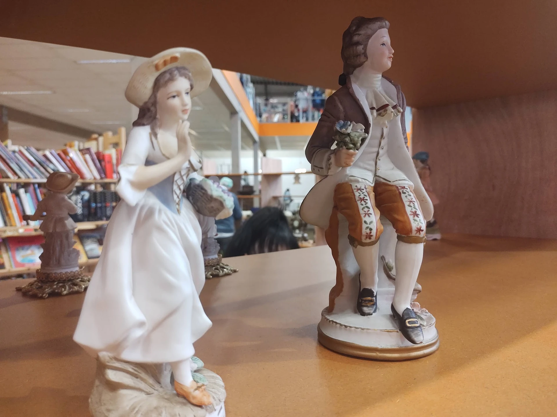 Twee decoratieve porseleinen beeldjes van een man en vrouw in historische kledij op een houten tafel in de winkel