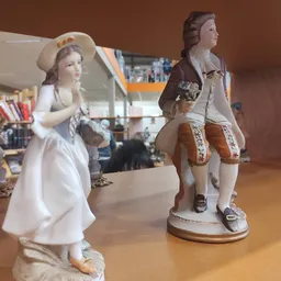 Twee decoratieve porseleinen beeldjes van een man en vrouw in historische kledij op een houten tafel in de winkel