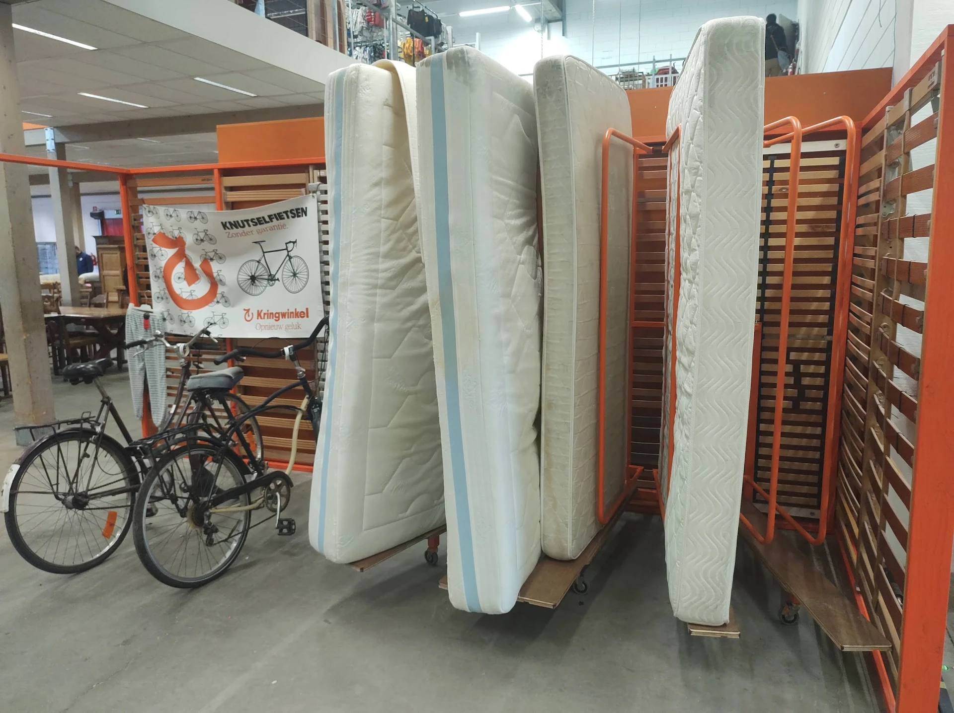 Matrassen en bedbodem uitgestald in de winkel, naast een fiets en oranje stellingen