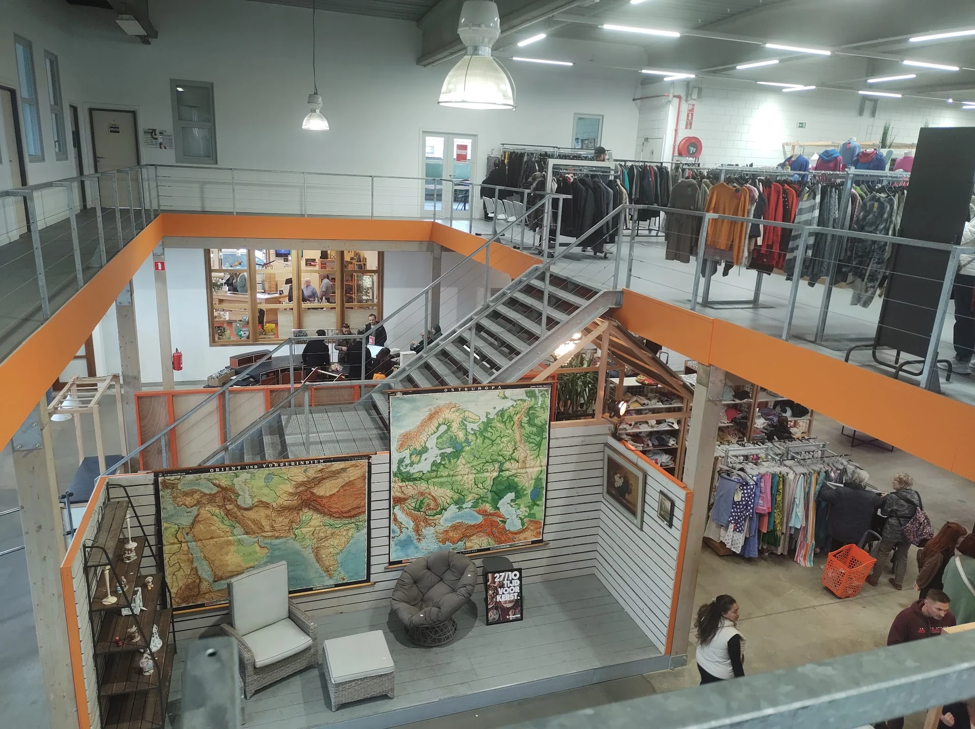 Overzicht van tweedehands winkel met oranje accenten, rekken met kleding op beide verdiepingen en themagebieden met decoratie