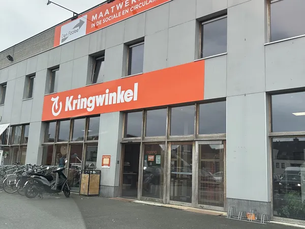 Gevel van kringloopwinkel met oranje Kringwinkel-logo en ingang, fietsen geparkeerd ervoor