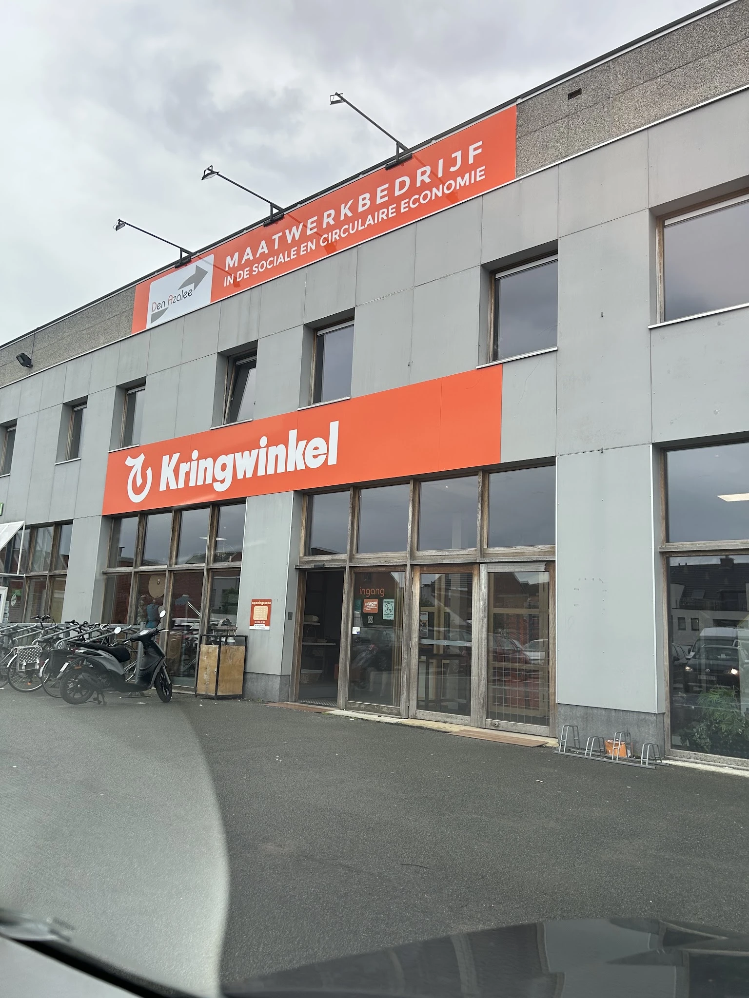 Gevel van kringloopwinkel met oranje Kringwinkel-logo en ingang, fietsen geparkeerd ervoor
