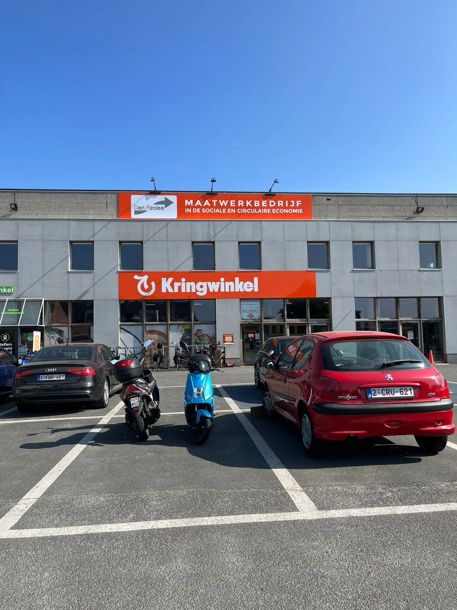Gevel van Kringwinkel met oranje letters, parkeerplaats met auto's en scooters ervoor