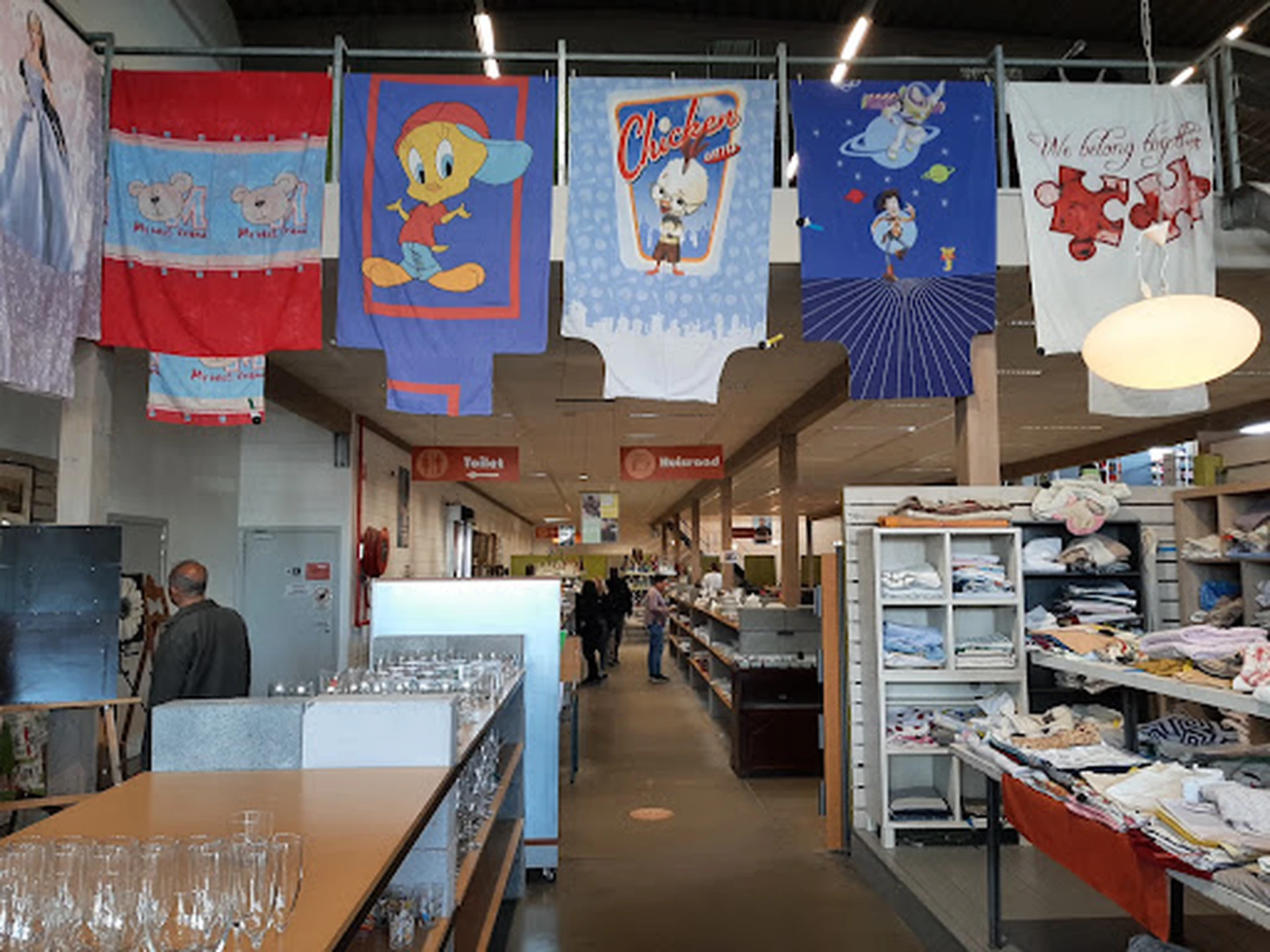 Binnenaanzicht van kringloopwinkel met gekleurde textiel aan touwen, rekken met tweedehandsartikelen en verlichte plafondlamp