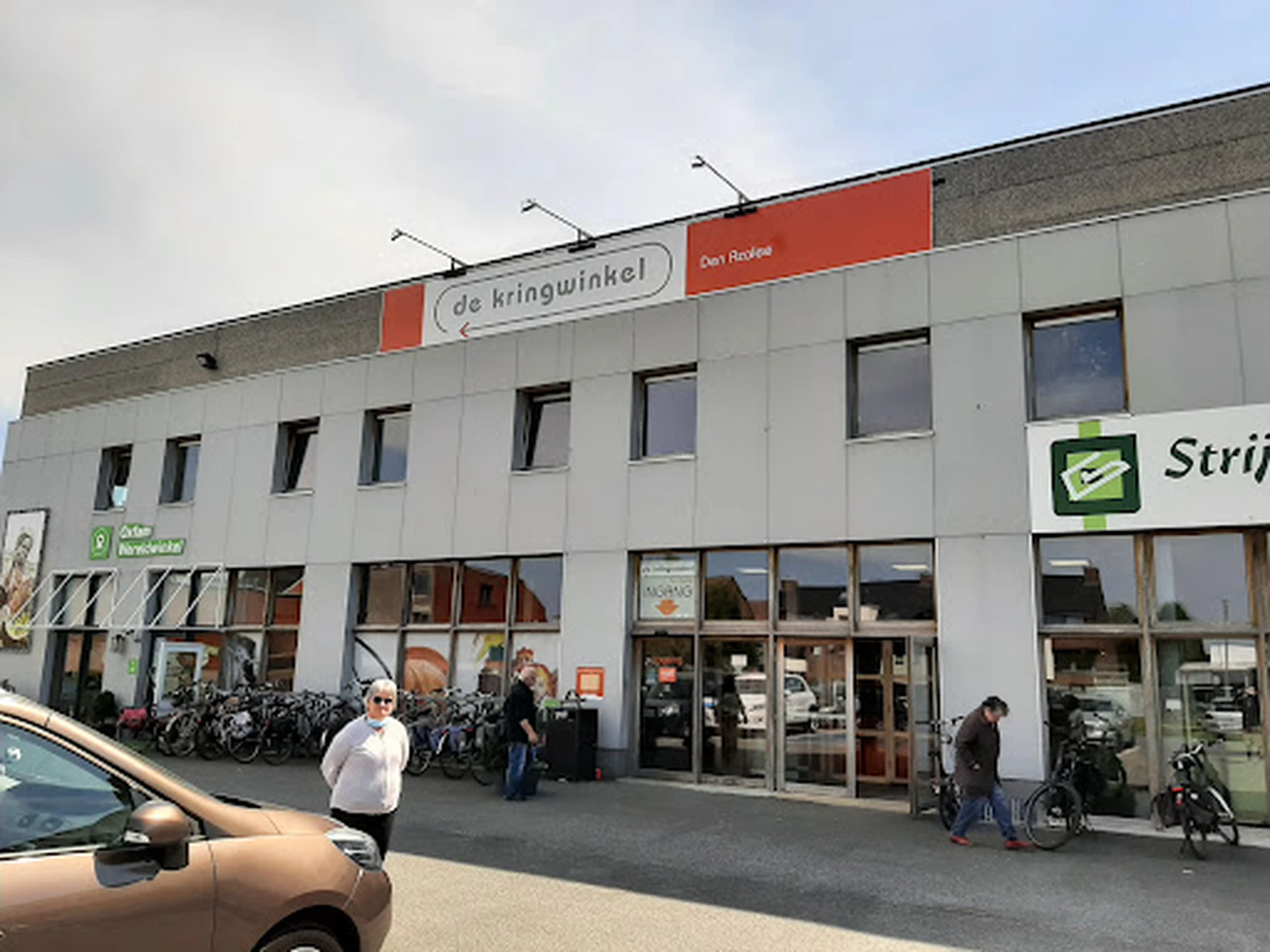Witte gevel van kringloopwinkel met rode signage, grote ramen en ingang met fietsen en bezoekers