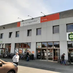 Witte gevel van kringloopwinkel met rode signage, grote ramen en ingang met fietsen en bezoekers