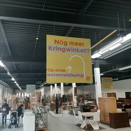 Geel hangend bord met Nederlandse tekst 'Nóg meer Kringwinkel?!' en 'Op onze bovenverdieping!' in een magazijnachtige ruimte