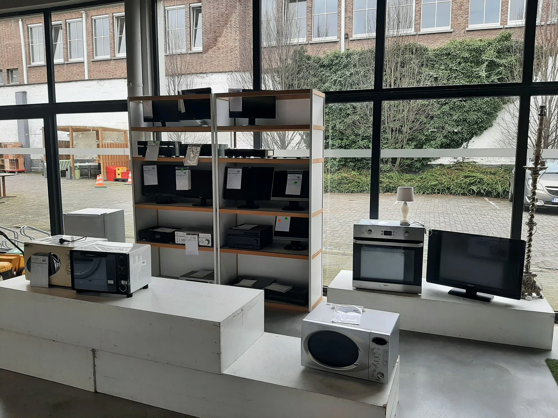 Uitgestalde tweedehands huishoudapparatuur: magnetrons, ovens en monitor op witte toonbanken
