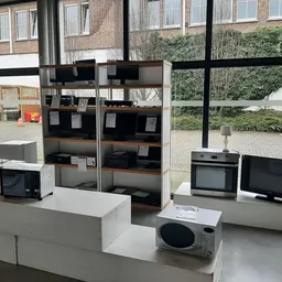 Uitgestalde tweedehands huishoudapparatuur: magnetrons, ovens en monitor op witte toonbanken
