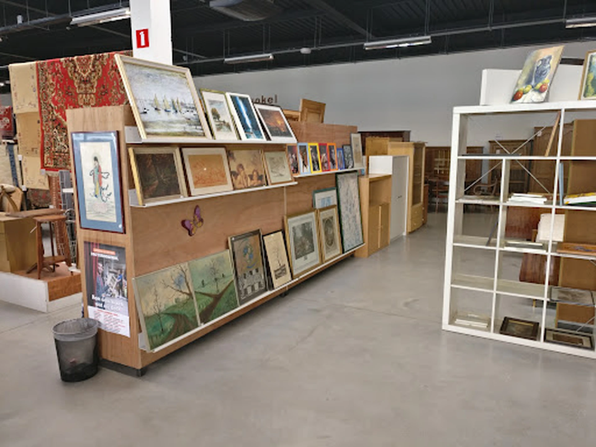 Houten stellingen en witte kasten gevuld met kunstwerken, schilderijen, prenten en boeken in een kringloopwinkel