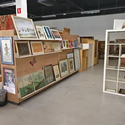 Houten stellingen en witte kasten gevuld met kunstwerken, schilderijen, prenten en boeken in een kringloopwinkel