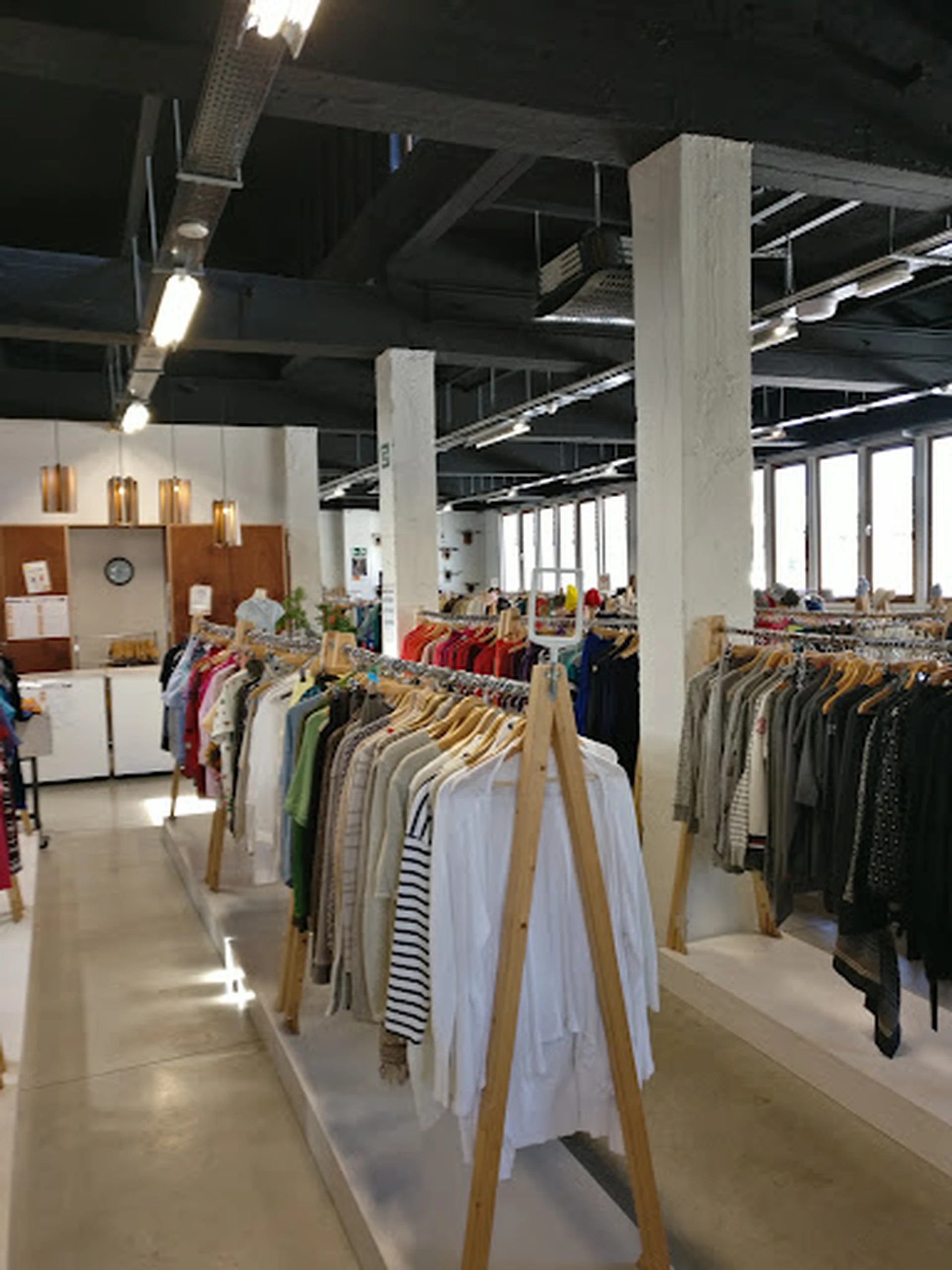 Ruim winkelinterieur met houten kledingrekken vol tweedehands kleding, grote ramen en industriële plafondstructuur