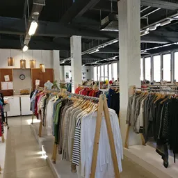 Ruim winkelinterieur met houten kledingrekken vol tweedehands kleding, grote ramen en industriële plafondstructuur