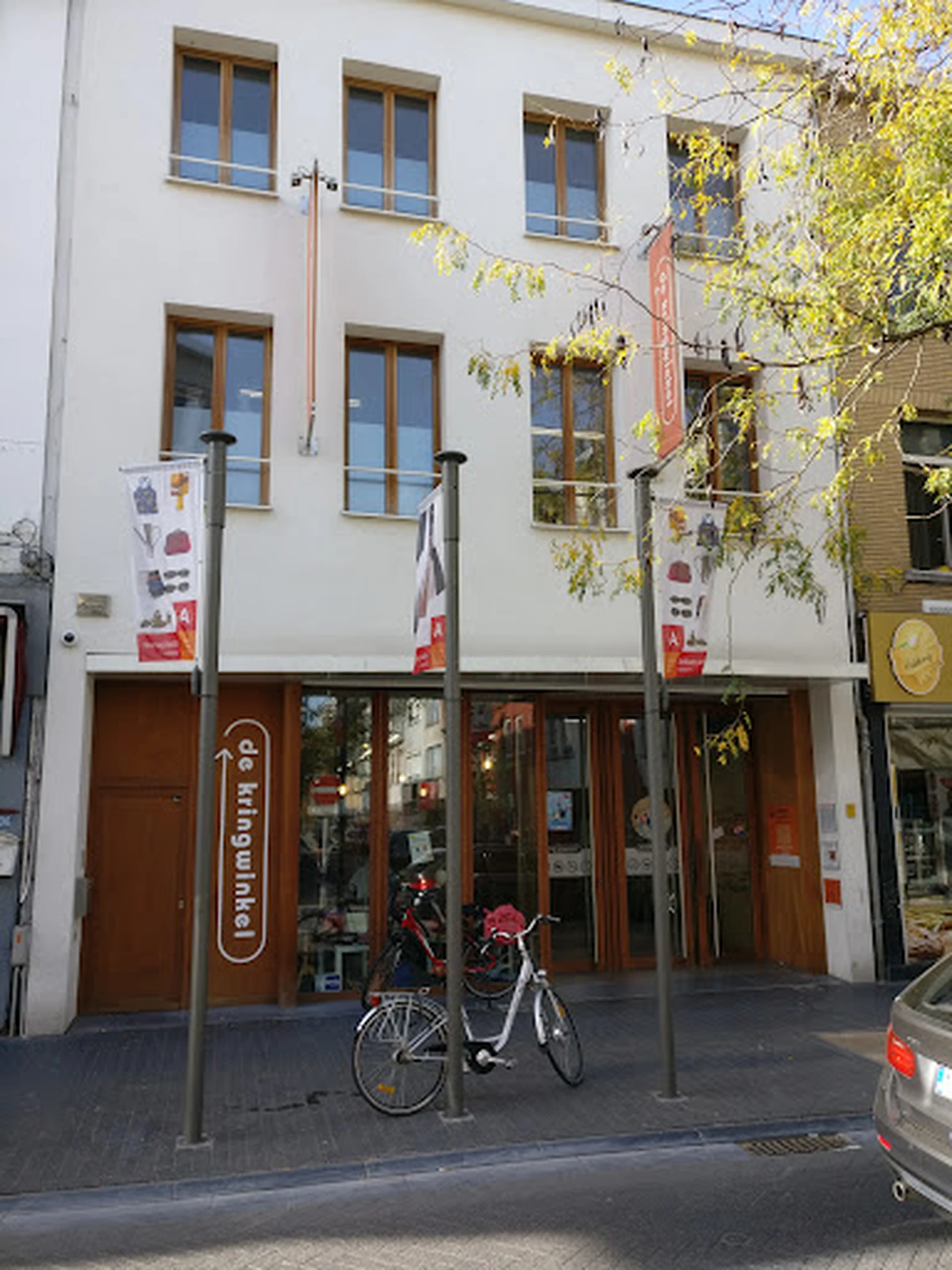 Witte gevel van kringloopwinkel met bruine deuren en ramen op begane grond, winkelnaam zichtbaar