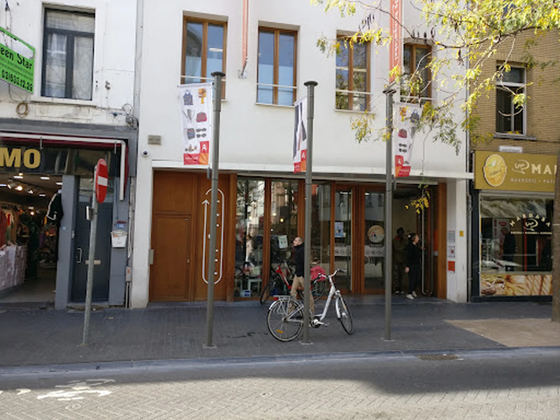 Gevel van kringloopwinkel met bruine deur, grote ramen en uithangsignage, fiets geparkeerd ervoor