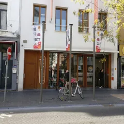Gevel van kringloopwinkel met bruine deur, grote ramen en uithangsignage, fiets geparkeerd ervoor