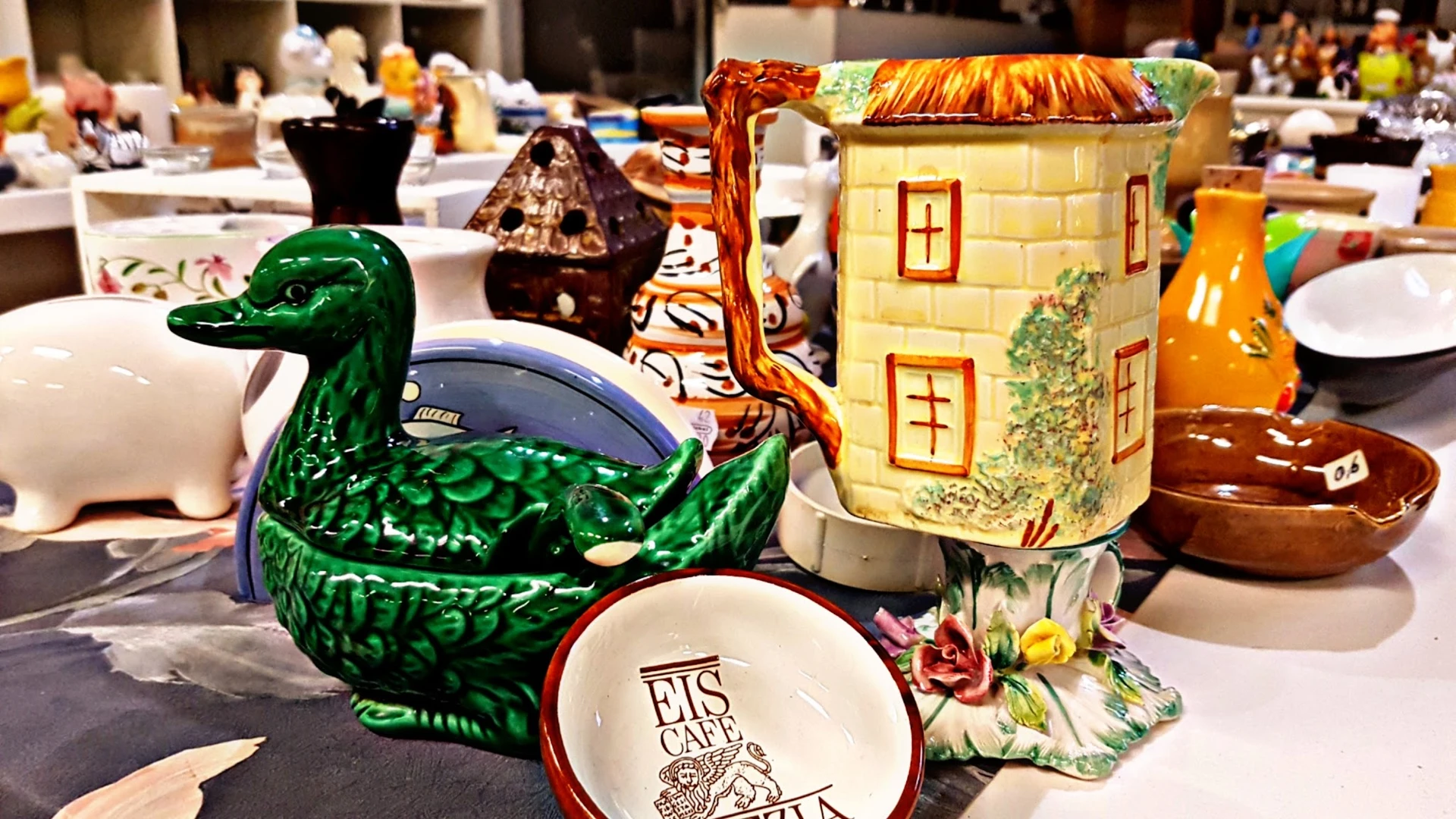 Verzameling decoratieve items op een tafel: groene eendenvaas, keramische huisjes, servies en andere vintage voorwerpen