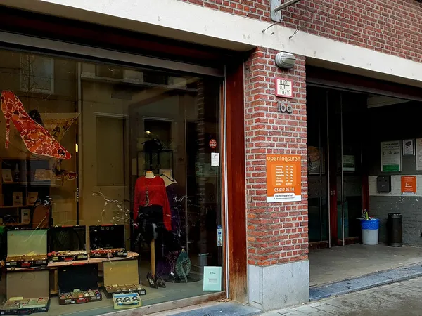 Voorgevel van kringloopwinkel met etalageruiten, rode deur, bakstenen gevel en oranje informatieborden