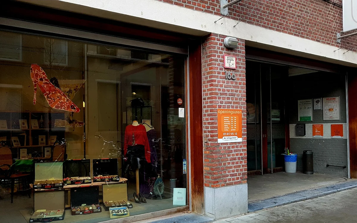 Voorgevel van kringloopwinkel met etalageruiten, rode deur, bakstenen gevel en oranje informatieborden