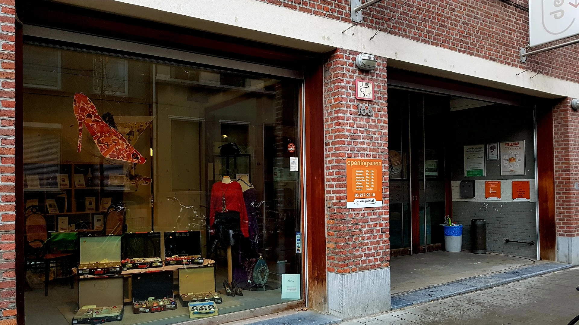 Voorgevel van kringloopwinkel met etalageruiten, rode deur, bakstenen gevel en oranje informatieborden