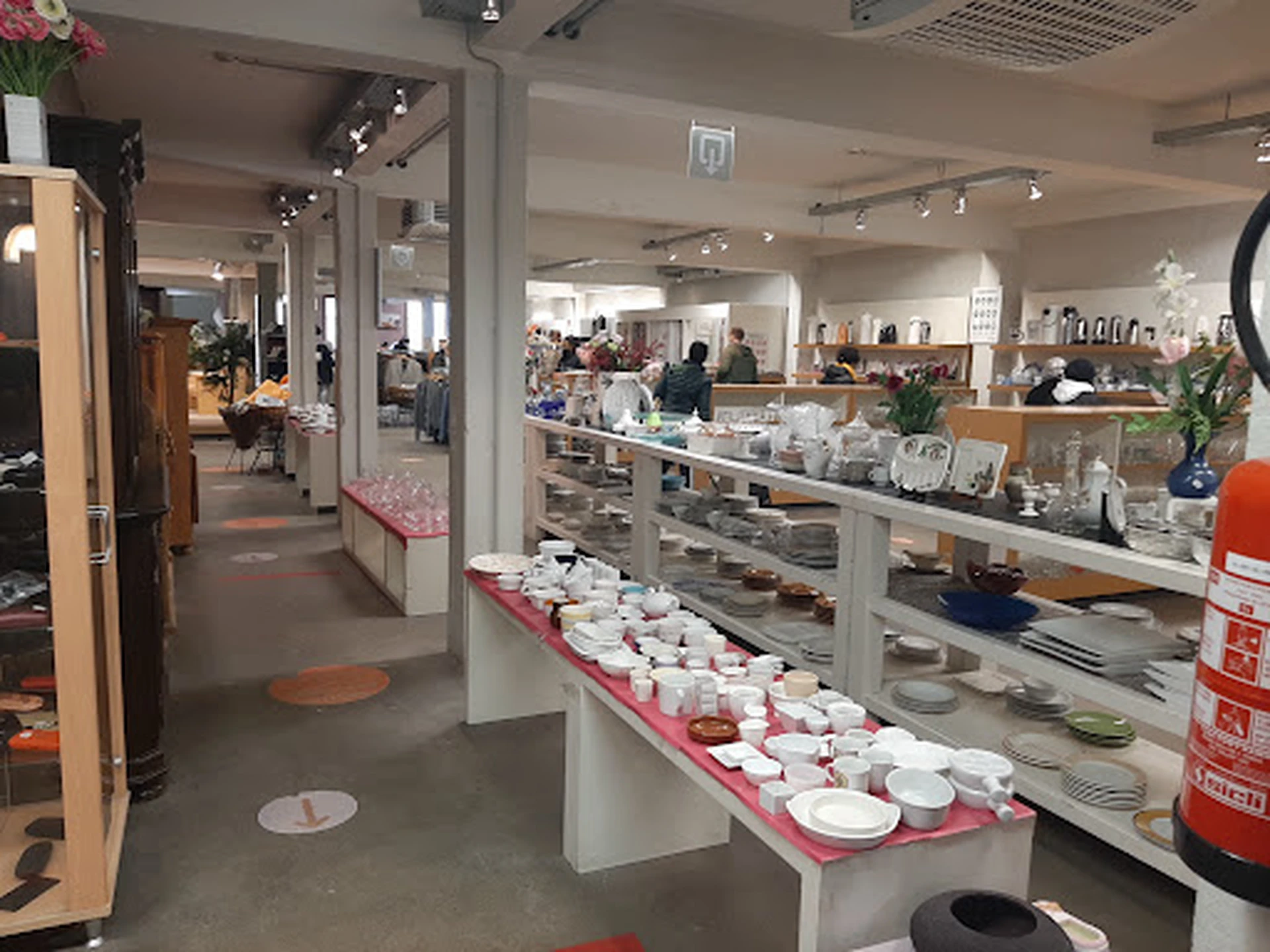 Winkelvloer met witte zuilen en roze displaykasten vol serviesgoed, klanten en stellingen op de achtergrond