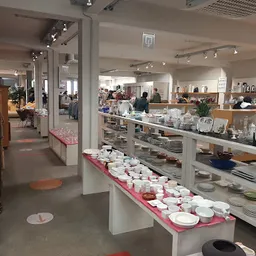Winkelvloer met witte zuilen en roze displaykasten vol serviesgoed, klanten en stellingen op de achtergrond