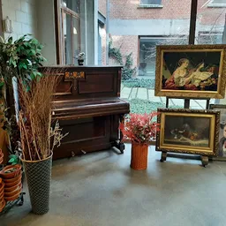 Winkelbinnenruimte met vleugelpiano, schilderijen op ezel, plantjes, oranje schalen en vintage decorstukken