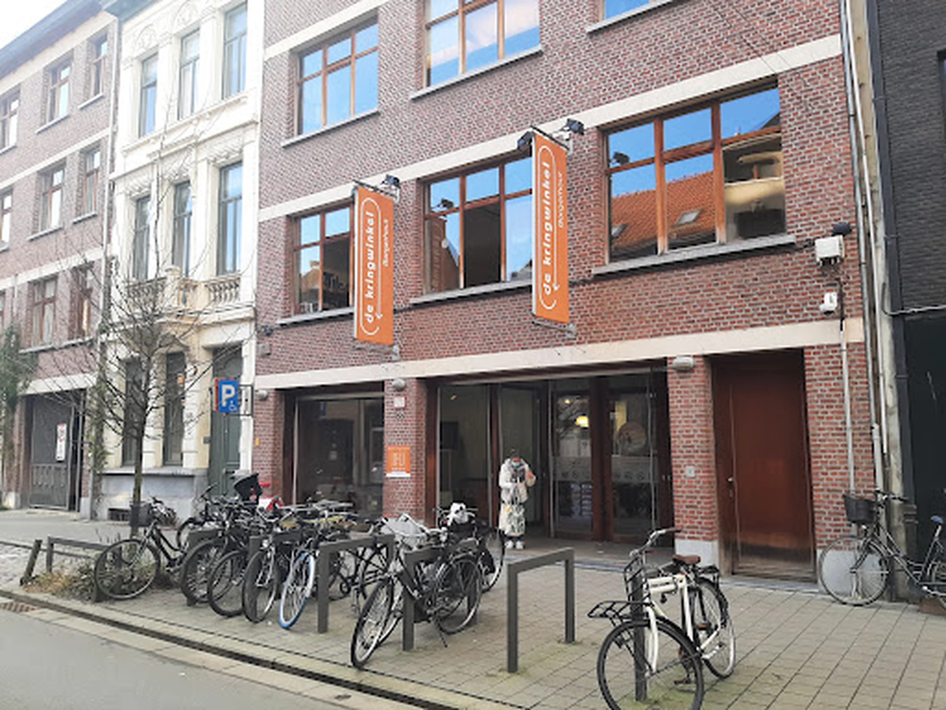 Bakstenen gevel van kringloopwinkel met grote ramen, oranje decoratieve elementen en fietsen geparkeerd voor de deur