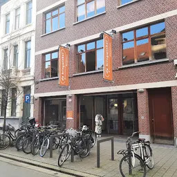 Bakstenen gevel van kringloopwinkel met grote ramen, oranje decoratieve elementen en fietsen geparkeerd voor de deur