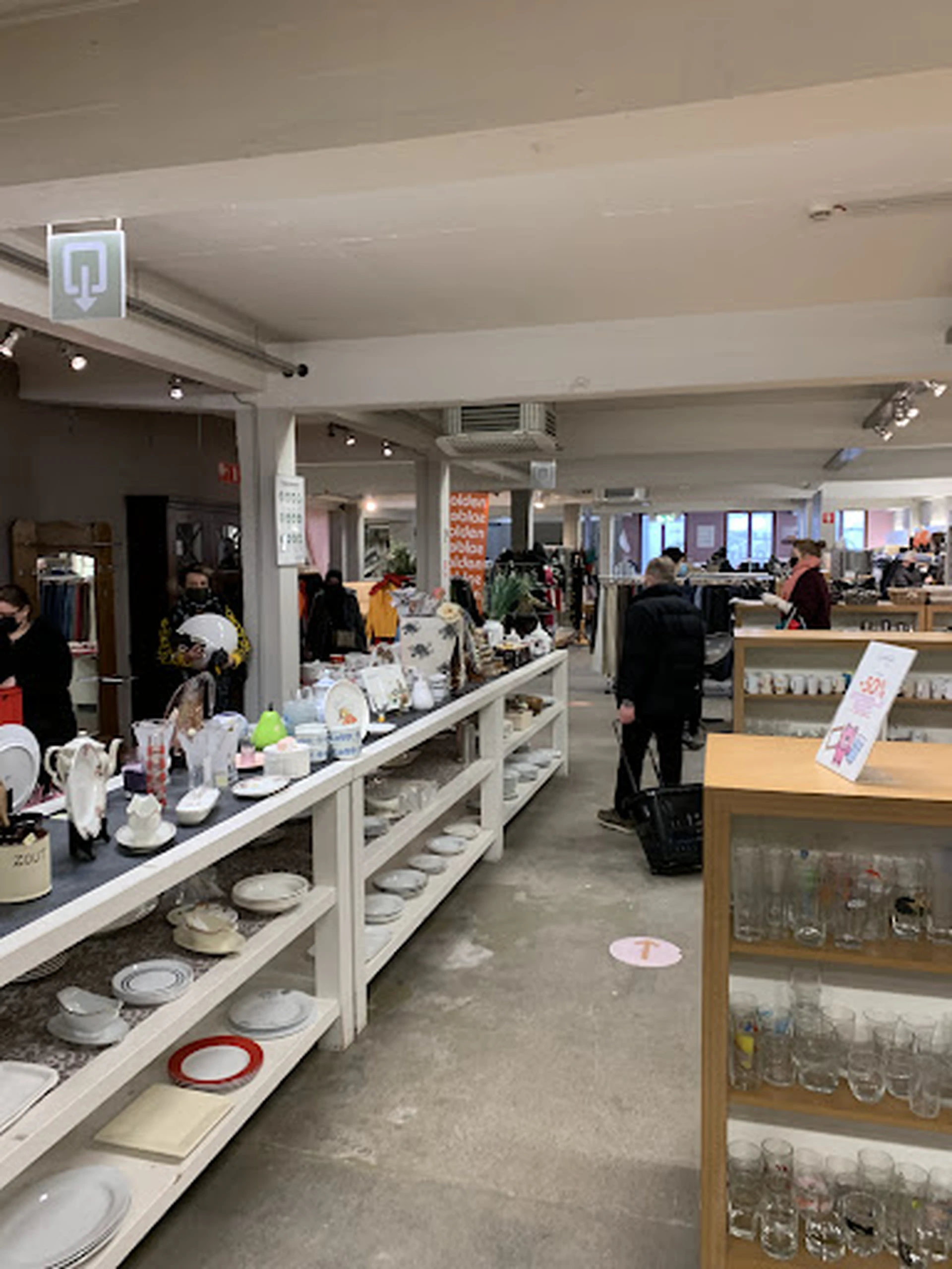 Winkelvloer met toonbanken vol serviesgoed, kandelaren en decoratie; klanten en stellingen zichtbaar in de winkel