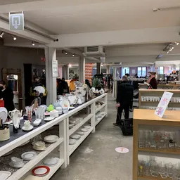 Winkelvloer met toonbanken vol serviesgoed, kandelaren en decoratie; klanten en stellingen zichtbaar in de winkel
