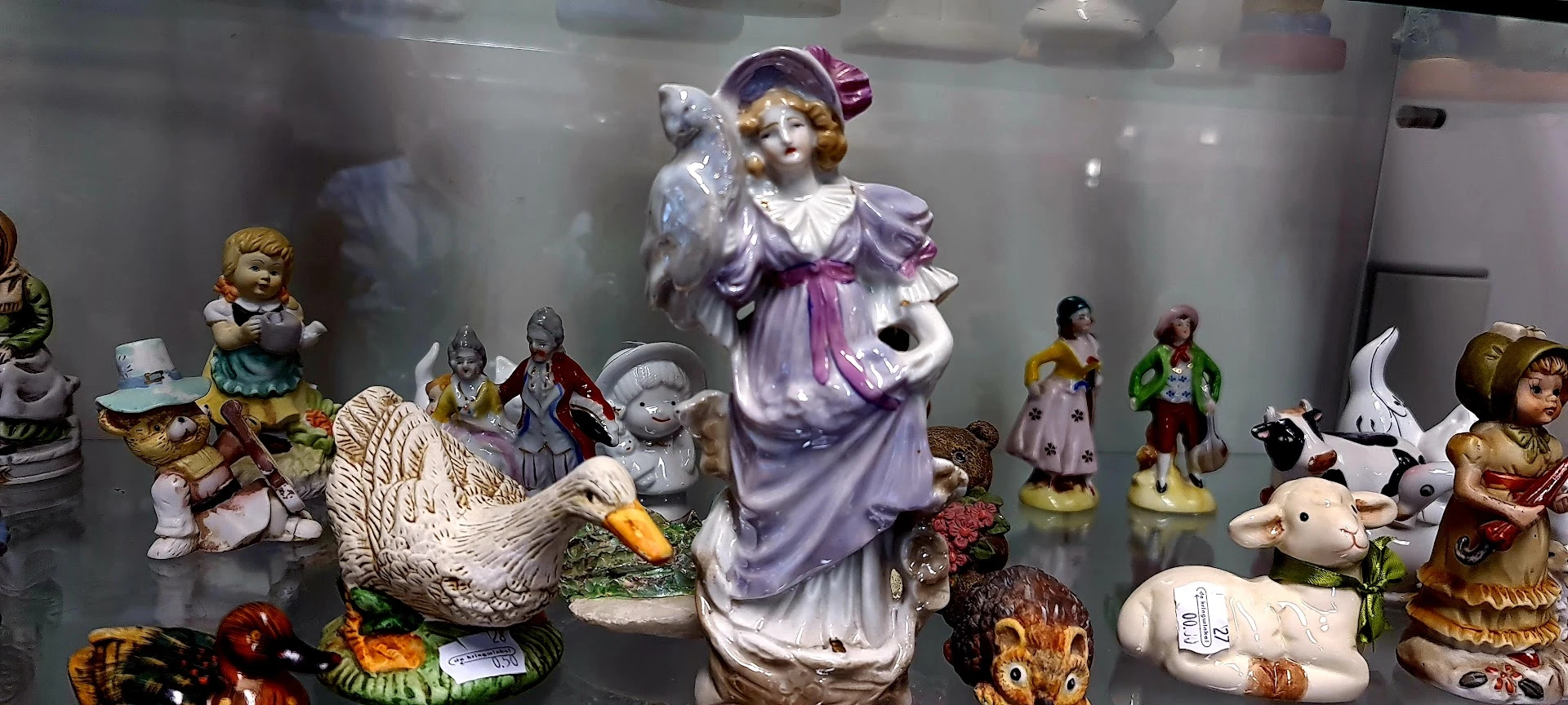 Collectie porseleinen beeldjes en figuren in een vitrinekasten, waaronder een vrouw in paarse jurk met bloemen en diverse die