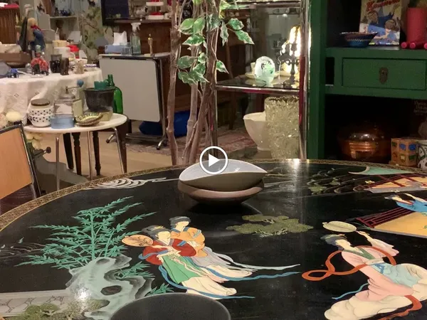 Werkruimte in kringloopwinkel met zwarte tafel met kunstwerk erop, potplant, glazenkast en diverse meubels en spullen op de a