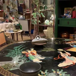 Werkruimte in kringloopwinkel met zwarte tafel met kunstwerk erop, potplant, glazenkast en diverse meubels en spullen op de a