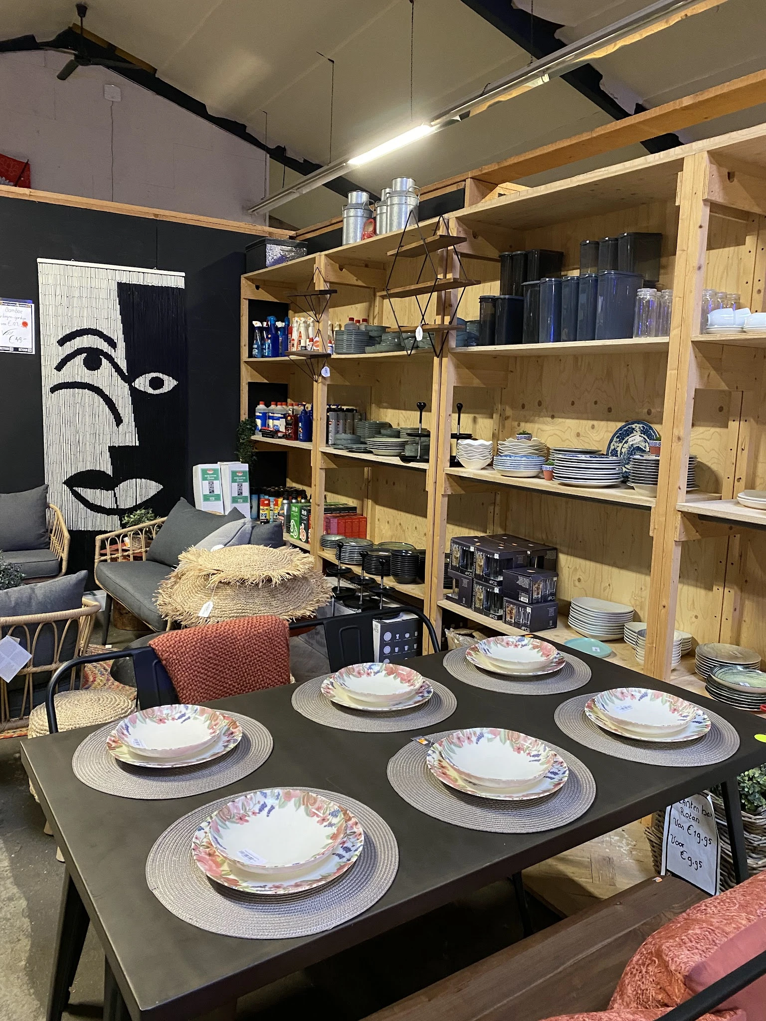 Winkelbinnenruimte met houten stellingen vol servies en huisraad, tafels met borden gedekt, kunstwerk aan muur