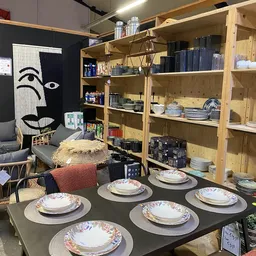 Winkelbinnenruimte met houten stellingen vol servies en huisraad, tafels met borden gedekt, kunstwerk aan muur