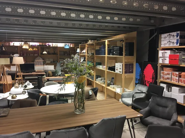 Winkelvloer van kringloopwinkel met houten tafel, zwarte stoelen, houten kast met spullen, planken met items en sfeervolle ve