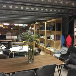 Winkelvloer van kringloopwinkel met houten tafel, zwarte stoelen, houten kast met spullen, planken met items en sfeervolle ve