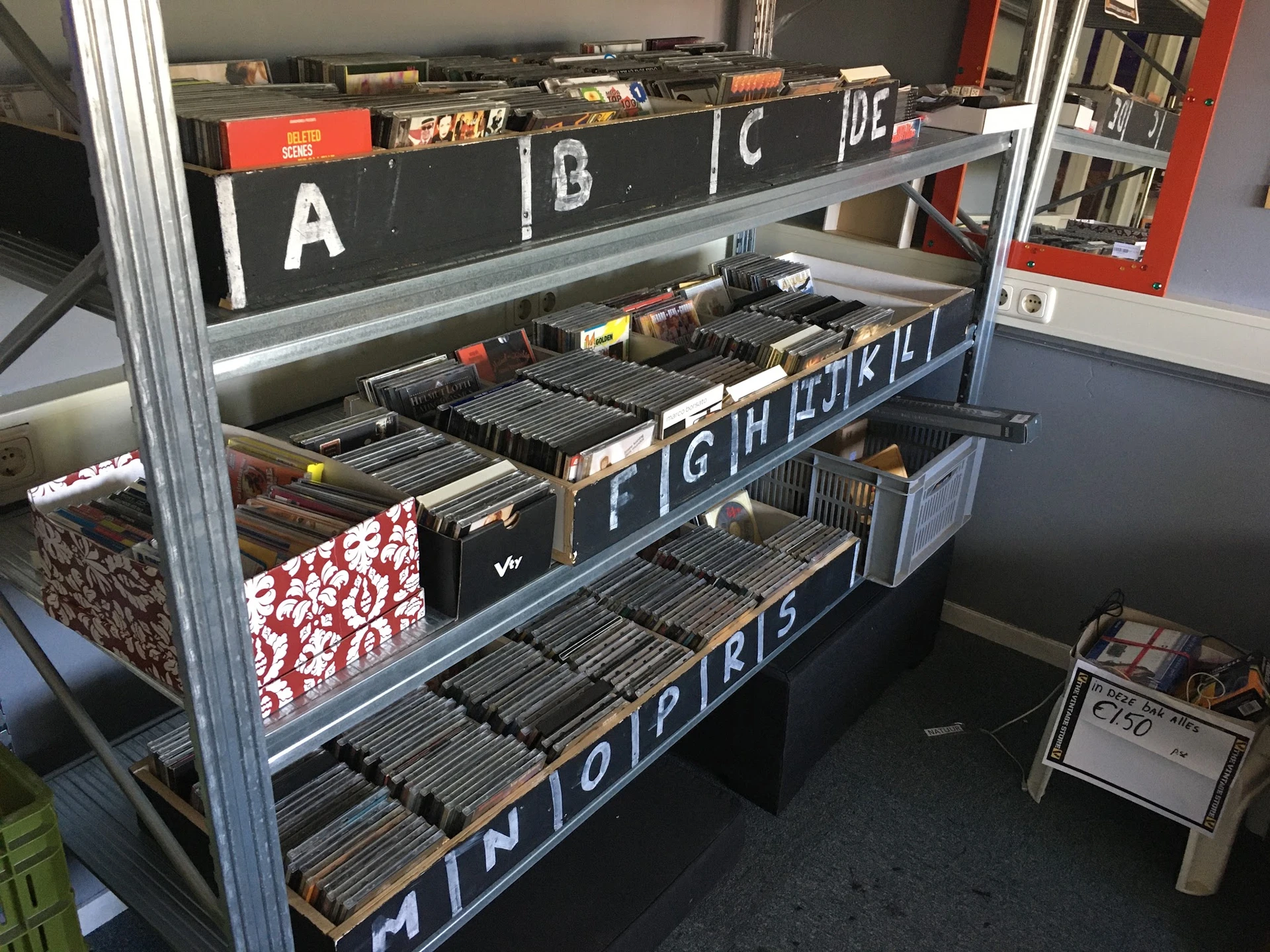 Metalen stellingkast met meerlaagse bakken gevuld met vinyl records en cd's, georganiseerd per letter