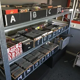 Metalen stellingkast met meerlaagse bakken gevuld met vinyl records en cd's, georganiseerd per letter