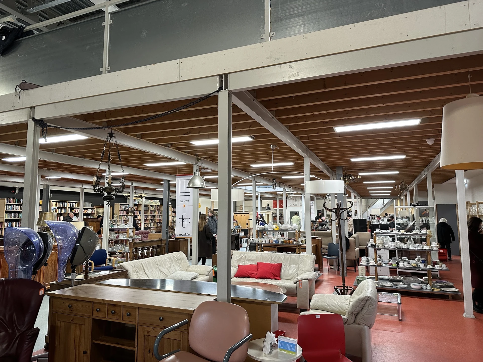 Ruim winkelinterieur met houten balkenplafond, stellingen met boeken, meubels en huishoudartikelen verspreid over de vloer