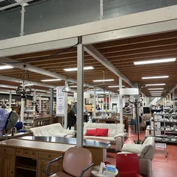 Ruim winkelinterieur met houten balkenplafond, stellingen met boeken, meubels en huishoudartikelen verspreid over de vloer