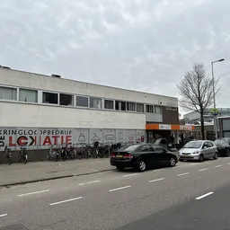 Voorgevel van kringloopwinkel met witte gevel, ramen en uithangtekst, parkeerplaats met auto's ervoor