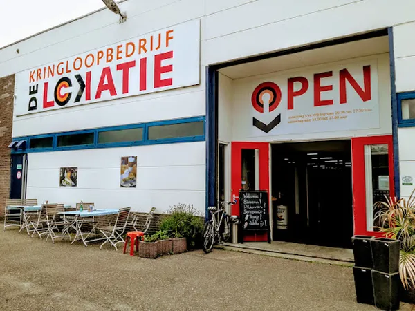 Voorgevel van kringloopwinkel met witte puien, rode deur en OPEN-bord, met terrasmeubilair op het plein ervoor
