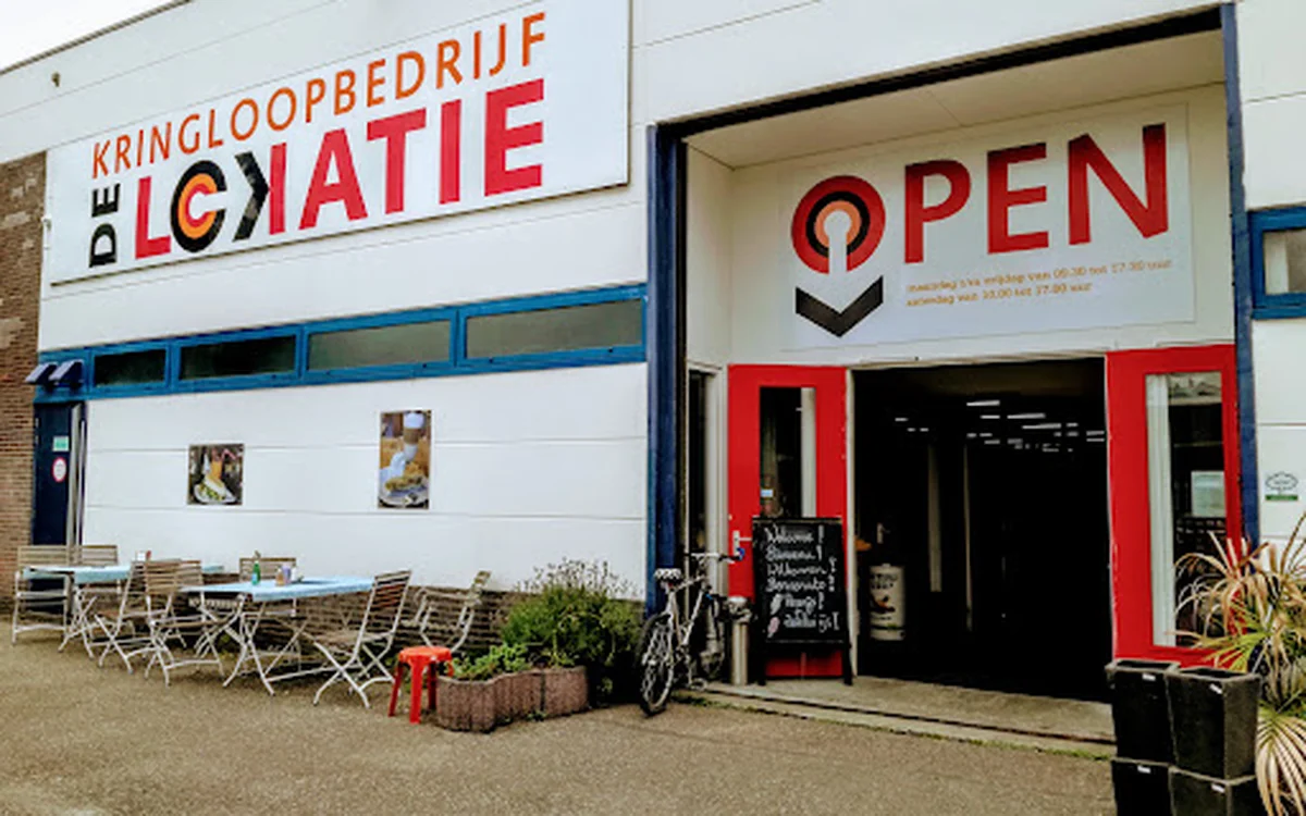 Voorgevel van kringloopwinkel met witte puien, rode deur en OPEN-bord, met terrasmeubilair op het plein ervoor