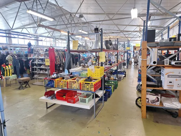 Winkelvloer met tafels vol spullen, kledingrekken, gele bakken en diverse tweedehandsartikelen onder industrieel plafond