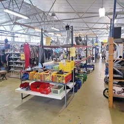 Winkelvloer met tafels vol spullen, kledingrekken, gele bakken en diverse tweedehandsartikelen onder industrieel plafond