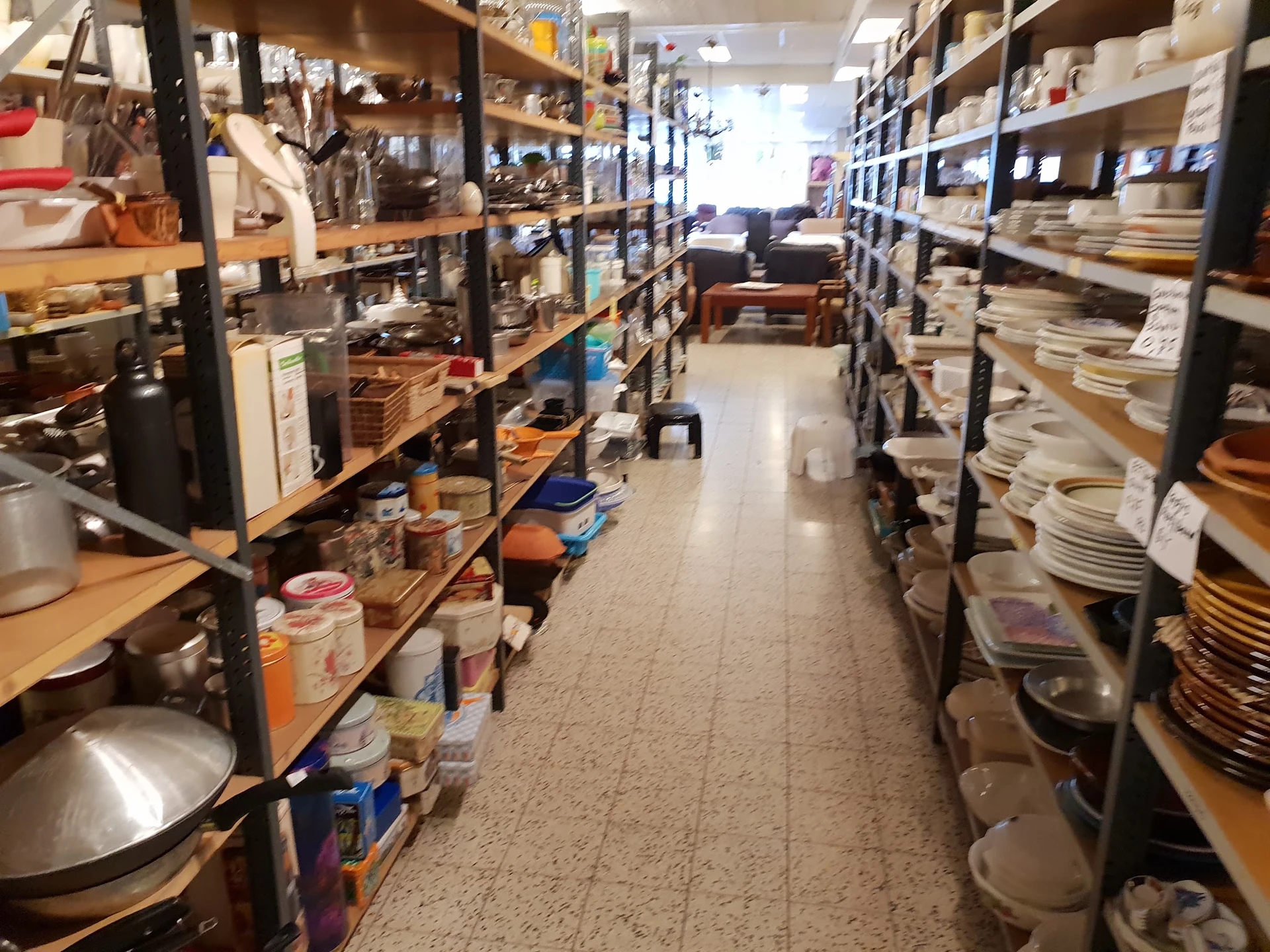 Lange winkelgang met metalen stellingen aan beide zijden volgestapeld met huishoudartikelen, servies en keukengerei