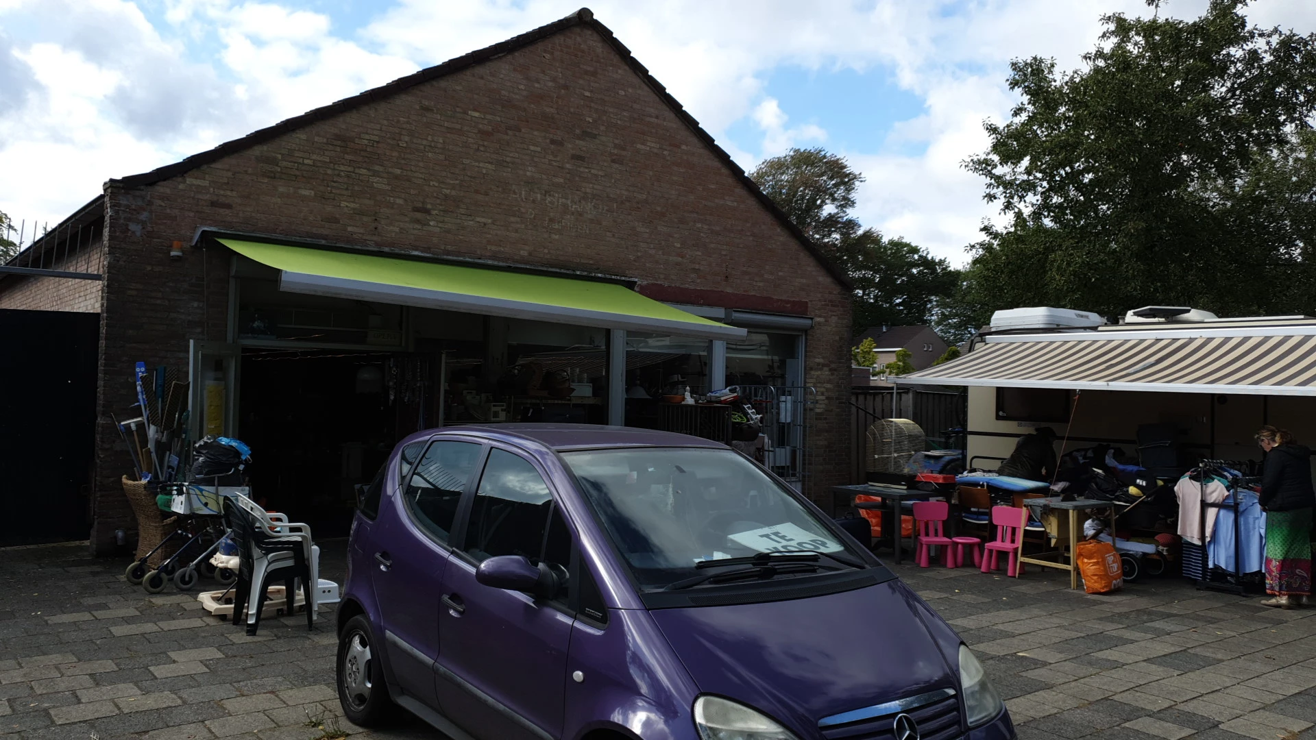 Voorkant kringloopwinkel met groene luifel en paarse auto op parkeerplaats
