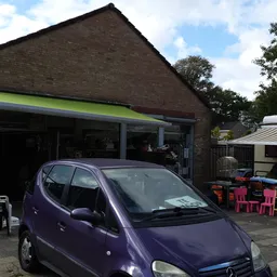 Voorkant kringloopwinkel met groene luifel en paarse auto op parkeerplaats